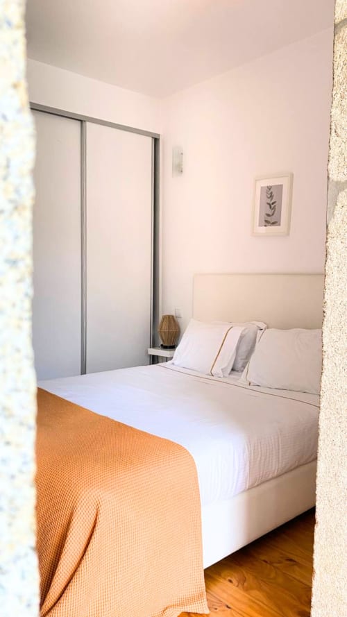 Quarto luminoso com cabeceira creme, roupa de cama branca, manta laranja, piso de madeira e guarda-roupa espelhado em Bom