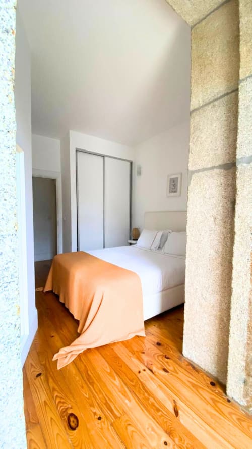 Quarto luminoso no Bom Porto Apartments com piso de madeira, cama branca com manta laranja, guarda-roupa espelhado e pedra bege