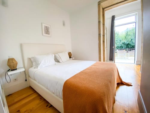 Quarto moderno nos Bom Porto Apartments no Porto com cama branca, manta laranja, assoalho em madeira e acesso a terraço com vista para o jardim