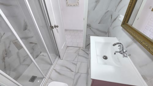 Baño moderno de mármol en VIVE Benalmádena con lavabo blanco, grifo cromado, ducha de cristal y marco dorado decorativo