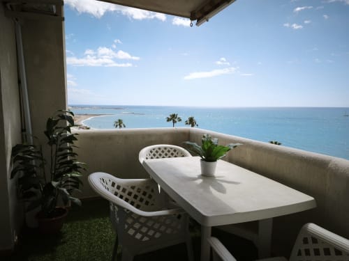 Terraza balcón en VIVE Benalmádena con mobiliario blanco, plantas en macetas y vistas panorámicas del Mar Mediterráneo con palmeras