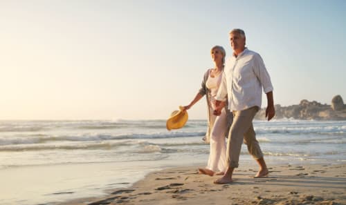 Pareja madura caminando descalza en la playa de Zahara al atardecer, llevando zapatos, con olas y paisaje costero visible