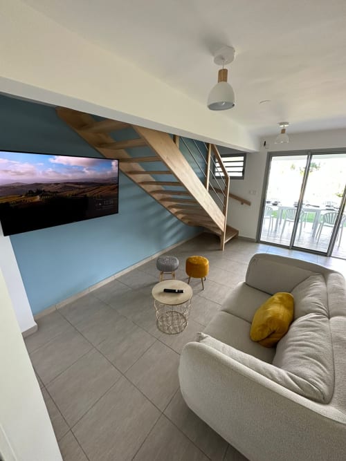 Élégant salon moderne à Maly Blue Location au Moule : canapé gris, escalier en bois, télévision et balcon avec vue sur l'océan