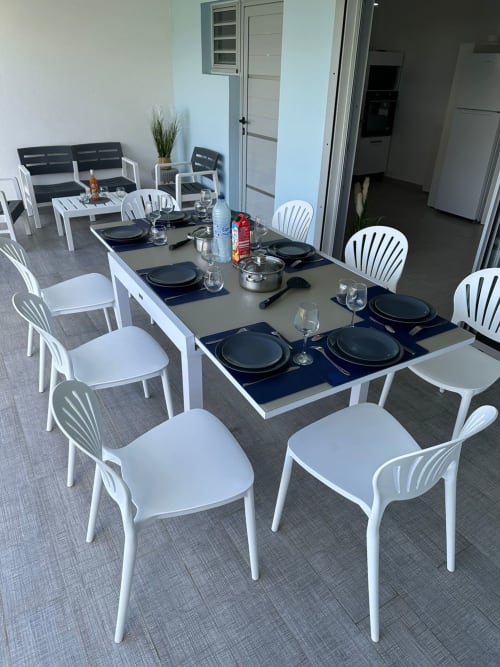 Élégante salle à manger à Maly Blue Location avec chaises blanches, table dressée aux assiettes noires et verres, cuisine visible