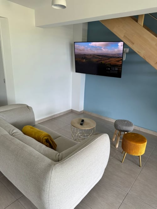 Élégant salon moderne au Maly Blue Location avec canapé courbe, téléviseur mural affichant un paysage, et poufs colorés
