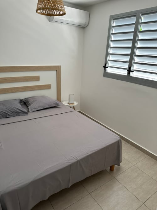 Chambre moderne à Maly Blue Location avec lit en bois, climatisation et fenêtres à persiennes avec vue sur mer au Locle