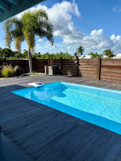 Piscine bleu vif sur terrasse en bois foncé avec palmiers et clôture en bois à Maly Blue Location, Le Moule, Guadeloupe