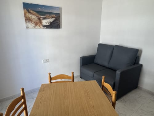 Sala de estar moderna en Apartamentos Playa Sol con mesa de comedor de madera, sofá negro y arte paisajístico costero en Es Canar