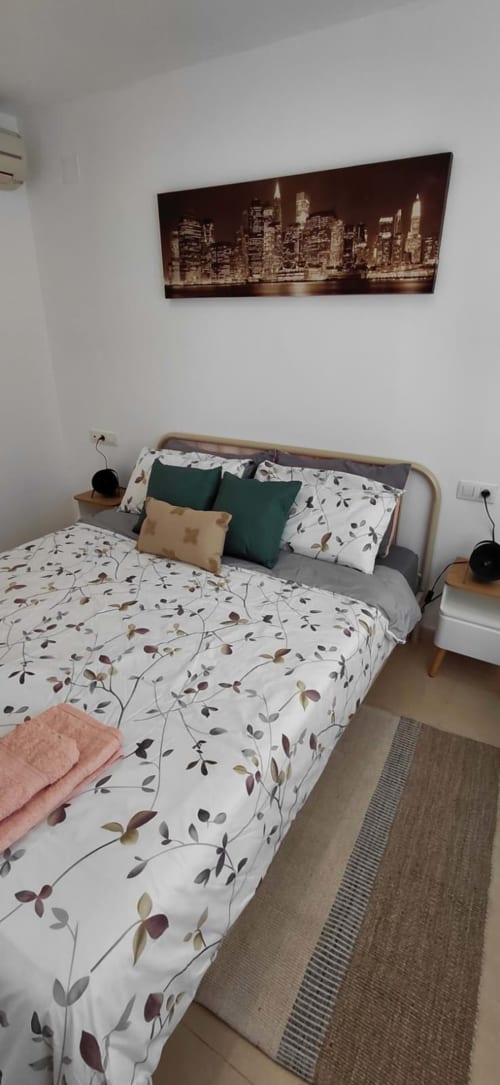 Chambre confortable dans un appartement de Benicàssim avec parure florale, tableau de skyline urbain, coussins verts et table de nuit en bois