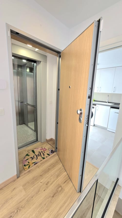 Open wooden apartment door with elevator entrance, hardwood flooring, and modern kitchen visible in García Sanguesa Sunny