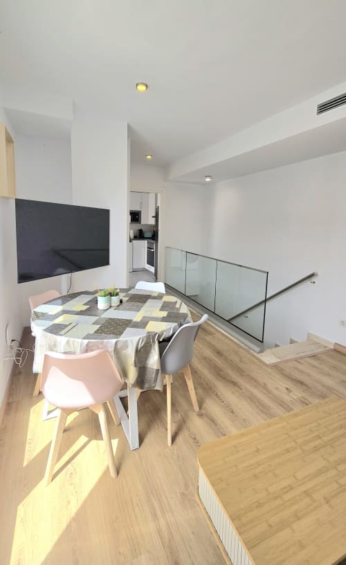 Modern apartment dining area with round table, pink chairs, wall-mounted TV, and glass railing overlooking kitchen at