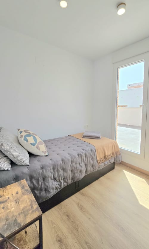 Bright modern bedroom in Benicàssim apartment with gray bed, wooden flooring, glass door opening to balcony with sea views,