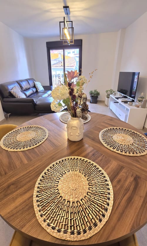 Modern dining area in Benicàssim apartment with wooden table, woven placemats, cream vase centerpiece, and city views through