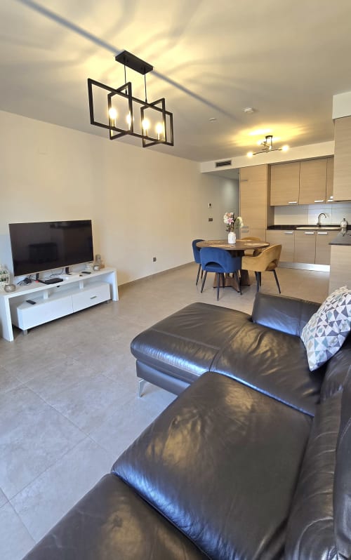 Modern apartment living room with black leather sofa, TV, dining area with blue chairs, and kitchen at Els Dolors Sunny Flat