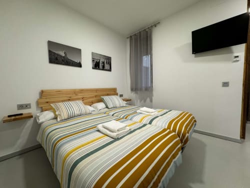 Habitación moderna en RJ26 San Isidoro León con cama de madera, ropa de cama a rayas, arte mural, TV y paredes blancas minimalistas