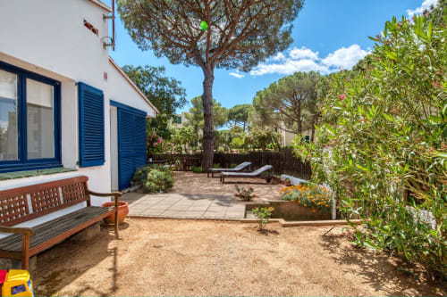 Jardí de caseta a Palafrugell amb terrassa, banc, gandules, arbres alts i casa blanca amb persianes blaves