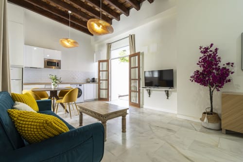 Modern apartment living room at G&H Apartments Jerez with blue sofa, yellow accents, kitchen, and purple flowering plant