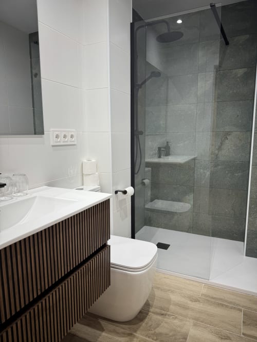 Bany modern a l'Hotel Oreneta de Gironella amb dutxa a ras de sòl, lavabo blanc i mobles amb aspecte de fusta