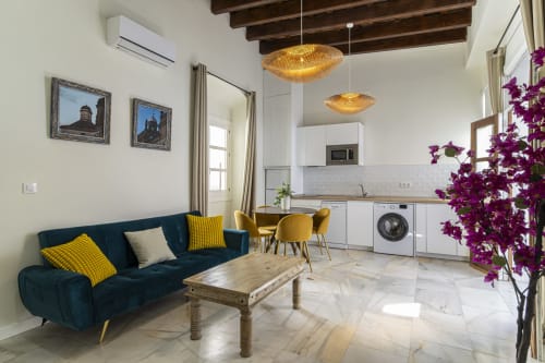 Modern apartment in Jerez de la Frontera with navy sofa, yellow accents, exposed beams, white kitchen, and purple flowers