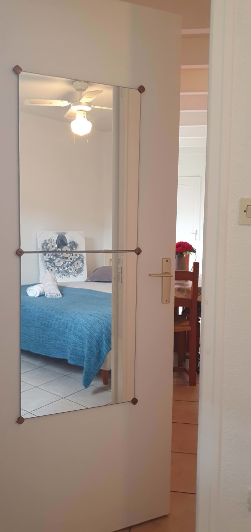 Vue du miroir de la chambre avec courtepointe bleue dans l'appartement de Carcassonne, ventilateur de plafond et couloir visibles