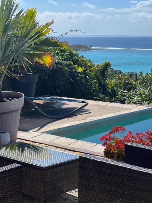Terrasse tropicale au bord de la piscine d'Aimehome avec vue sur l'océan, chaises longues et fleurs rouges vives surplombant les eaux turquoise
