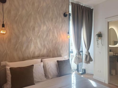 Camera da letto di moderno appartamento milanese con testata rivestita in beige strutturato, tende grigie, luci a sospensione e bagno ensuite in vista