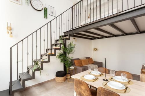 Zona living e pranzo in loft moderno con scala, tavolo per quattro, divano con cuscini arancioni e piante verdi da A Casa di