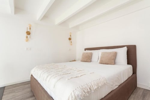 Camera da letto moderna e luminosa a Casa di Imma Milano con letto marrone, biancheria bianca, soffitto con travi e applique dorate