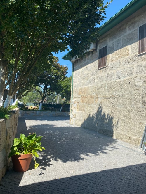 Pátio ensolarado da pousada Casa Augusta em Braga, Portugal, com edifício de pedra, árvores frondosas e plantas em vasos