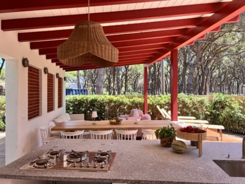 Casa Pineta's covered outdoor dining area with red beamed ceiling, gas cooktop, wooden table, and tree-lined garden view in