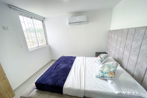Dormitorio moderno con cabecero tapizado gris, ropa de cama blanca, aire acondicionado y ventana en Olamar Living, Santiago de Tolú