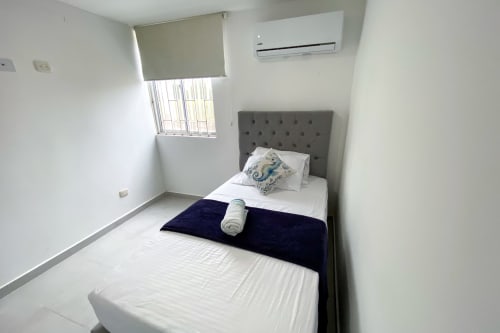 Habitación moderna en Olamar Living hotel en Santiago de Tolú con cabecera gris, ropa de cama blanca, aire acondicionado y ventana con persiana