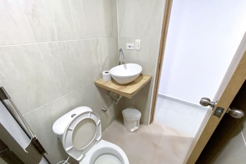 Baño moderno en apartamento hotel Olamar Living con inodoro blanco, lavamanos de cerámica sobre tocador de madera y paredes con azulejos beige