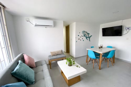 Sala de estar moderna con sofá gris, mesa de comedor con sillas azules, TV y arte de tiburón en pared del hotel Olamar Living