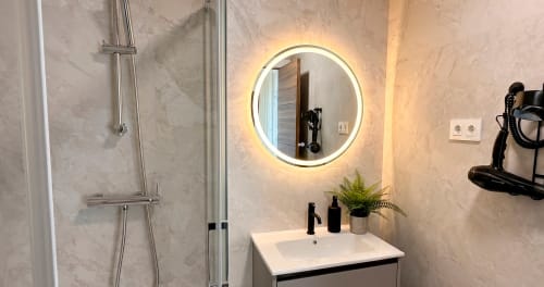 Baño moderno en La Posada de las Flores con espejo redondo iluminado, azulejos de mármol, lavamanos blanco y ducha walk-in