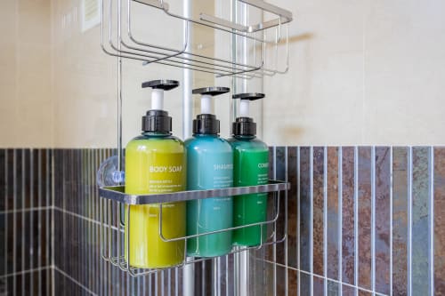 Chrome shower caddy with yellow body soap, turquoise shampoo, and green conditioner bottles in modern bathroom at Haus