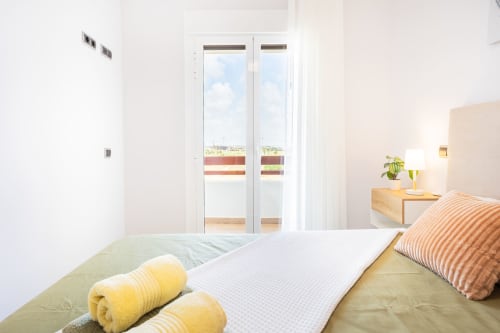 Bright bedroom with double bed, yellow towels, striped pillows, and glass doors opening to sea view balcony at Haus Holiday