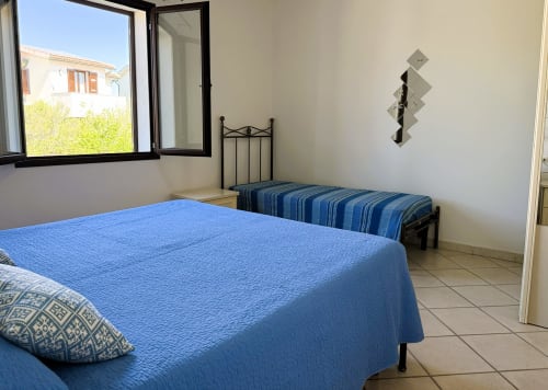 Camera luminosa con copriletto blu, secondo letto e finestra sul giardino della villa A Mare Case Vacanze a Valledoria
