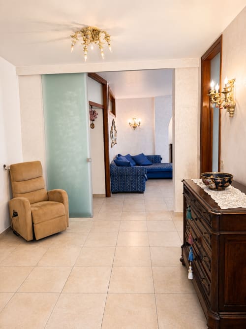Ampia zona living dell'Appartamento Lisa con poltrona reclinabile beige, divano blu, cassettone in legno e porte scorrevoli verso la camera.
