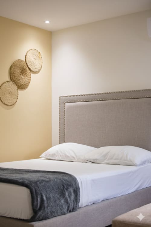 Dormitorio moderno en Olamar Living con cabecera tapizada, ropa de cama blanca, cestas tejidas decorativas en la pared y tonos amarillo suave