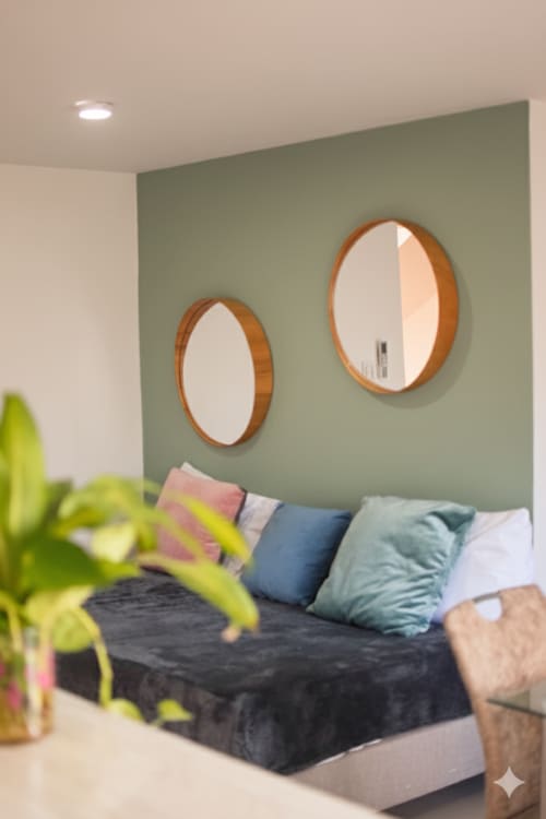 Habitación moderna en Olamar Living con pared de acento verde salvia, dos espejos redondos de madera, almohadas coloridas en cama oscura, y