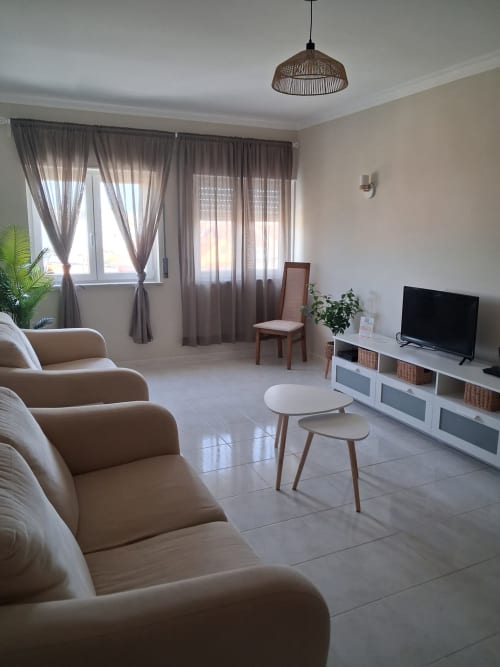 Helles modernes Wohnzimmer in der Furnaka Village Herberge in Lourinhã mit beigefarbenen Sesseln, Fernseher, weißen Möbeln und grauen Vorhängen