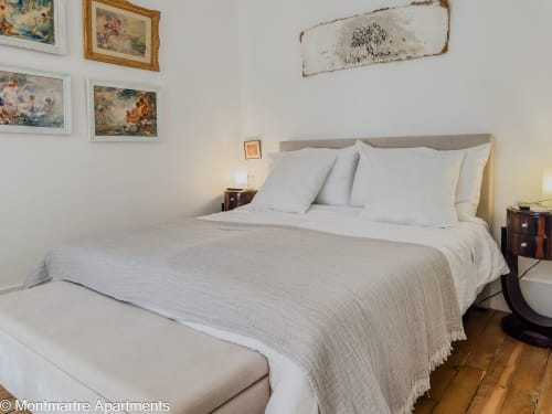 Elegant bedroom in Montmartre Apartments Paris with white bedding, gray headboard, framed artwork, and wooden flooring.