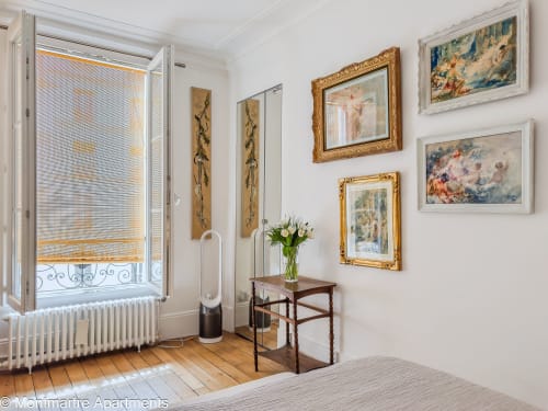 Bright Parisian apartment bedroom with artistic gallery walls, wooden desk with fresh flowers, and Eiffel Tower balcony view