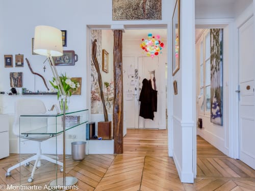 Modern Paris apartment workspace with glass desk, white chair, wooden beams, colorful floral art, and open-concept living