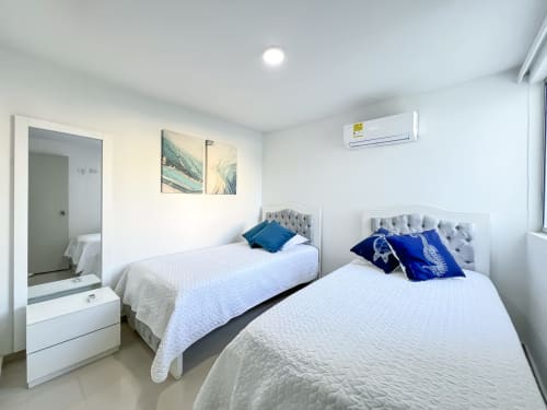 Dormitorio luminoso con dos camas individuales, ropa de cama blanca, almohadas azules, cabeceros grises, aire acondicionado y decoración moderna en Mar