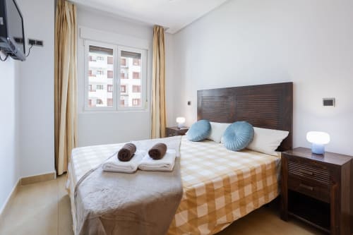 Dormitorio moderno en apartamento Haus Holiday Homes en Los Alcázares con cama de madera, almohadas azules y vistas a la ciudad
