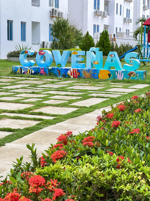 Colorido letrero 'COVEÑAS' con arte de peces tropicales en el jardín del hotel Olamar Living, con flores rojas y jardín manicurado