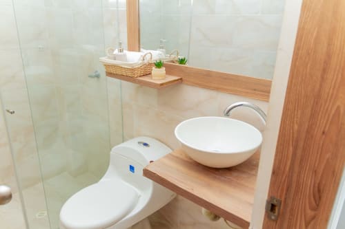 Baño moderno con inodoro blanco, lavamanos redondo, tocador de madera y ducha con mamparas de vidrio en Olamar Living, Coveñas