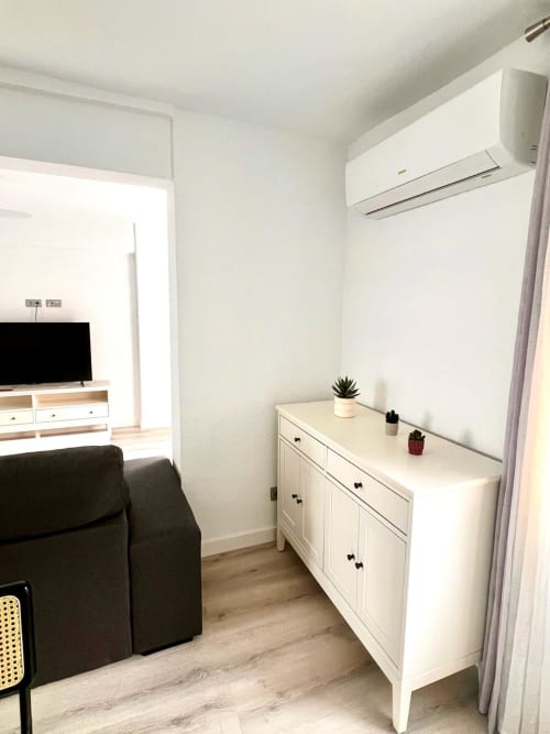 Moderno dormitorio de apartamento con cómoda blanca, silla oscura, pisos de madera y aire acondicionado mural en Águilas